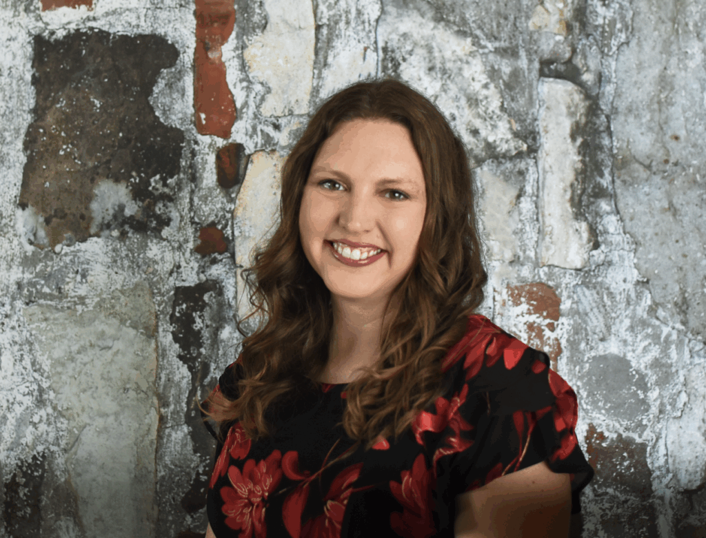 A woman with long wavy brown hair, wearing a black dress with red floral patterns, smiles in front of a rustic, weathered stone and brick wall.